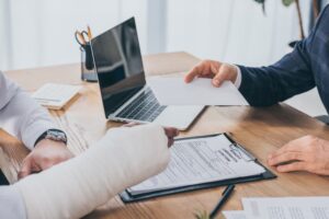 cropped view of businessman sitting at table and giving paper sheet to worker with broken arm in office, compensation concept